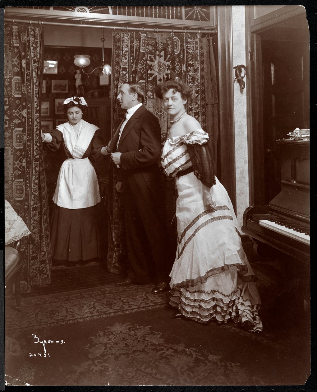 正式な服装の男性と女性、ピアノのある部屋で家事のように見えるもの、1905年（銀ゼラチンプリント） 作： バイロンカンパニー