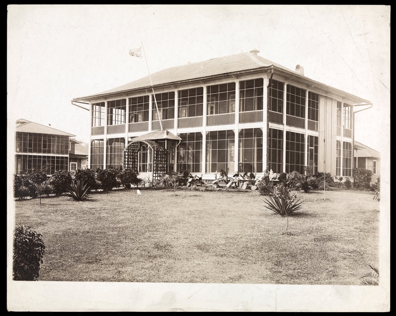 ポーチに囲まれた2階建ての家で、アメリカの旗が飛んでいます。 1912年に運河の建設中にパナマで（銀ゼラチンプリント） 作： バイロンカンパニー
