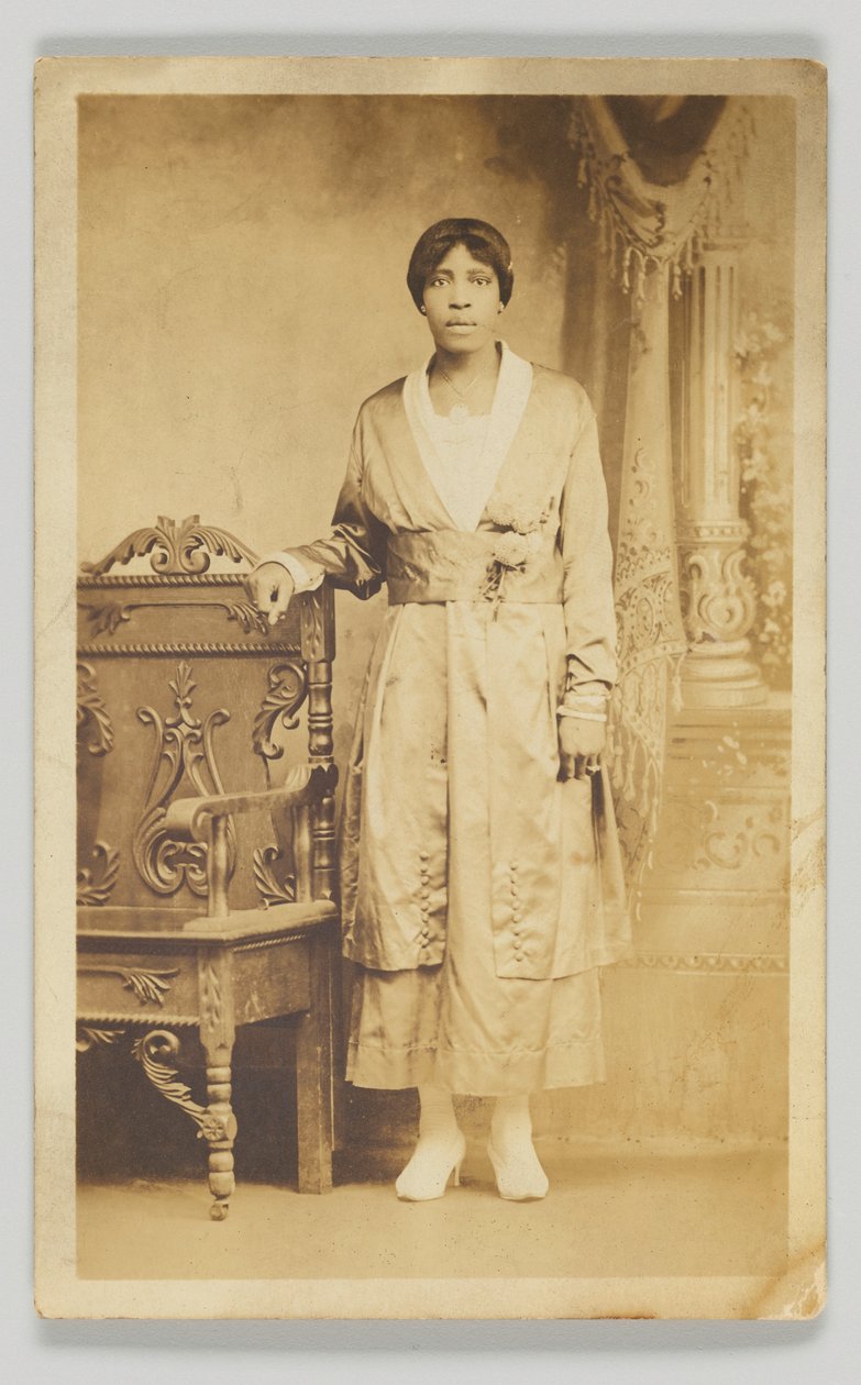 木製の椅子の隣に立つ女性の写真はがき、1904～1918年。 作： 不明