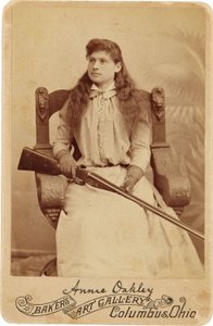 アニー・オークリー : Annie Oakley:.ルネッサンス様式の巨大な椅子に控えめに座り、ショットガンを膝の上に置く伝説の狙撃手。彼女の繊細で女性的な特徴と流れるような髪は、彼女の頑丈なアウトドアイメージを裏切る ©Buyenlarge/UIG/Leemage 作： American Photographer