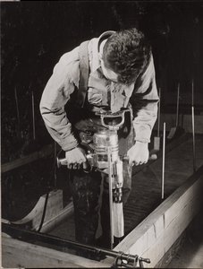 [男は手持ちの機械で木材を加工する]。 作： Arnold Eagle