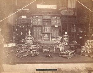 S.ハワード＆サンズ社の展示--本館、布張りの椅子、テーブル、暖炉、キャビネット、シェルフユニットなどの家具のコレクション。壁には絨毯、テーブルや棚には花瓶が置かれています。 作： Centennial Photographic Co.