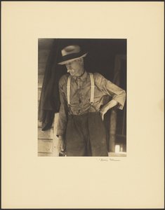 [麦わら帽子をかぶり、サスペンダーをつけた髭面の老人が戸口に立っている] 作： Doris Ulmann