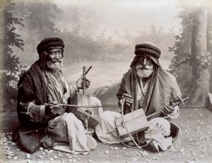 控えめな民族衣装を着た2人のアラブのミュージシャンの肖像画。年配の男性は、地面に座ってスタジオでポーズをとっています。それらの後ろには、森の風景の背景があります 作： Félix Bonfils