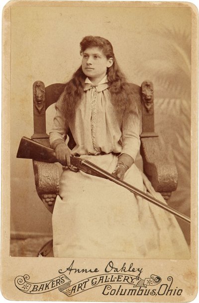 アニー・オークリー : Annie Oakley:.ルネッサンス様式の巨大な椅子に控えめに座り、ショットガンを膝の上に置く伝説の狙撃手。彼女の繊細で女性的な特徴と流れるような髪は、彼女の頑丈なアウトドアイメージを裏切る ©Buyenlarge/UIG/Leemage 作： American Photographer