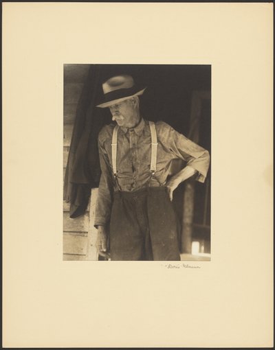 [麦わら帽子をかぶり、サスペンダーをつけた髭面の老人が戸口に立っている] 作： Doris Ulmann