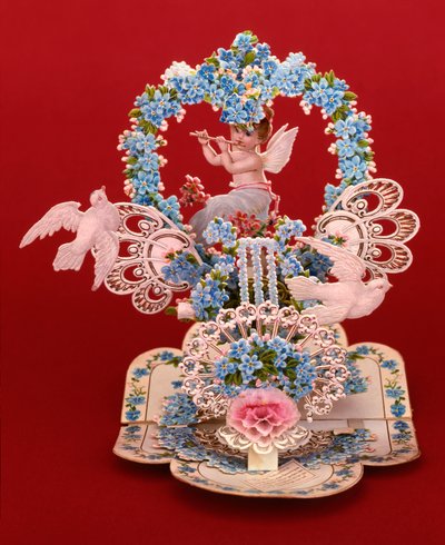 忘れな草のハート型アーチの下で、翼のあるケルブがフルートを吹いているポップアップカード（クロモリトグラフ・カード 作： English School