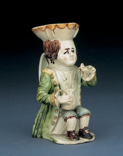 トッパーのトービージャグ、茶色と黄色のスカラップで縁取られた帽子、切り刻まれた茶色の髪が並んでいて、白いロゼットが膝に手すり子の水差しを置いて座っている、c.1770 作： English School