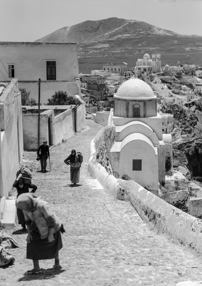 白壁の建物が並ぶギリシャの村の石畳の道を歩く人々。 作： Maynard Owen Williams