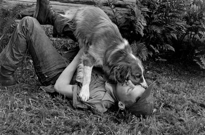 アラスカ州パーマーのログハウスの外で犬と遊ぶ青年 作： Ted Spiegel