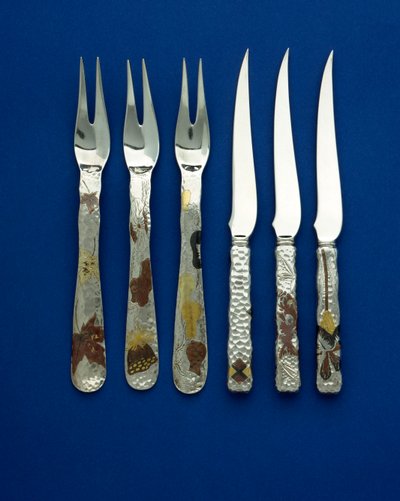 36本のデザートナイフとフォークのセットの一部。ハンマーで叩かれたハンドルには、パティーヌ加工された銅と変色した金がはめ込まれており、様々な花や動物、幾何学的なデザインの詳細が彫られています。 作： Tiffany and Company