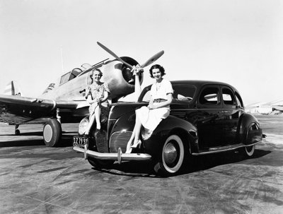 California: 1939 空港で1939年型リンカーン・ゼファー・セダンのフェンダーに座る2人の女性、その後ろには単発の飛行機が停まっている（モノクロ写真）。 作： Unbekannt
