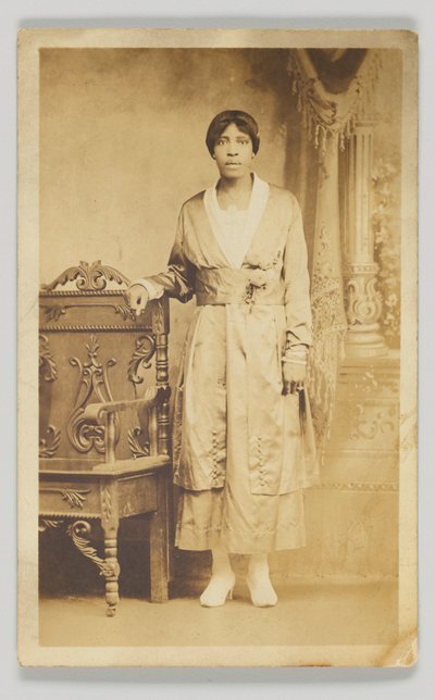 木製の椅子の隣に立つ女性の写真はがき、1904～1918年。 作： Unbekannt