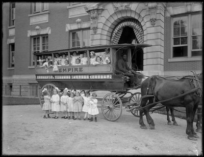 小さな女の子が馬車の前と前で、正体不明の建物の前でポーズをとっています。 作： William Davis Hassler