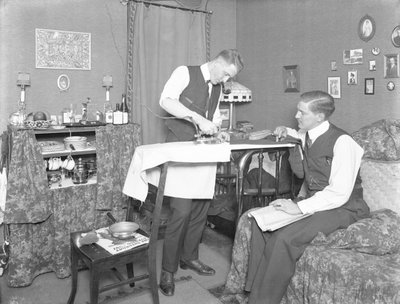 電気アイロンの使用を示す正体不明の男性で、別の男性が見守っています。 作： William Davis Hassler