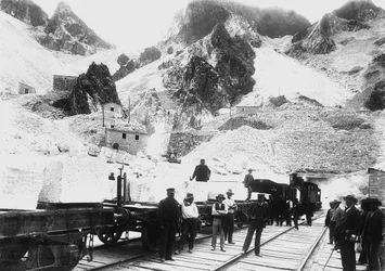 アプアンアルプスのラヴァッチョーネで、大理石の塊を積んだ鉄道車列の前に立つ掘削機(モノクロ写真)