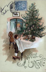 クリスマス・グリーティングカード：テーブルの上に置かれたクリスマスツリーの前でポーズをとる子供と、窓から見える天使 - 1905年のポストカード